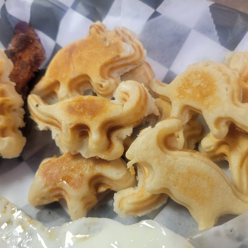 Dinosaur-shaped waffles with a side of fried chicken and a dollop of white sauce.