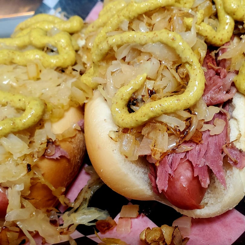 Two hot dogs with mustard, sauerkraut, and pastrami on a checkered and pink paper.