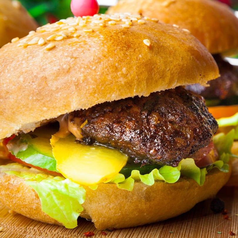 Close-up of a juicy burger with a sesame seed bun, lettuce, pickles, and tomato.