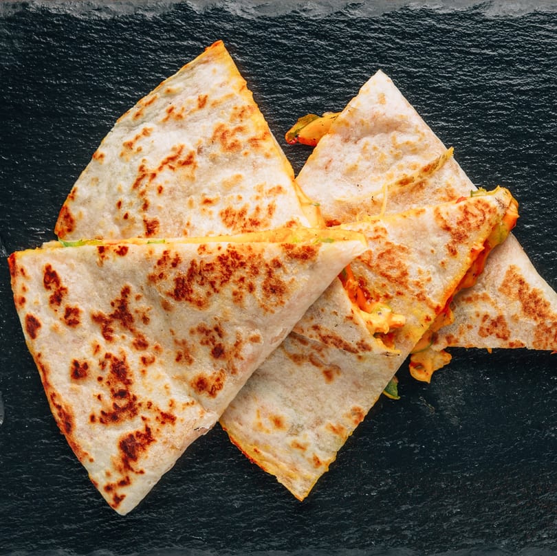 Four triangular quesadilla slices with melted cheese and vegetables on a dark textured surface.